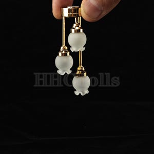 Puede incluir: Una lámpara de araña de oro en miniatura con tres bombillas de vidrio esmerilado. La lámpara de araña cuelga de una cadena de oro.