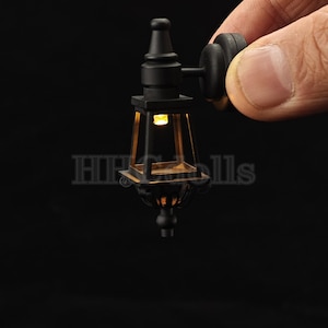 May include: A miniature black metal wall-mounted lantern with a clear glass panel and a warm white LED light. The lantern has a decorative design and is perfect for dollhouses or miniature scenes.