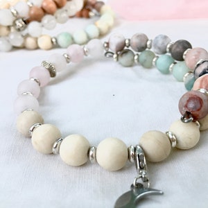May include: A bracelet made of natural stone beads in cream, pink, grey, and brown hues. The bracelet has silver-coloured metal accents and a crescent moon charm. Other bracelets are visible in the background.