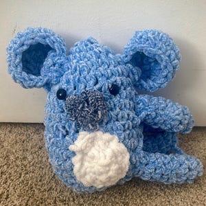 May include: A blue crocheted koala plush toy with white fur on its belly and black eyes.