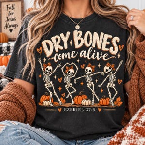 May include: A charcoal gray t-shirt featuring a graphic print with the text "DRY BONES come alive" and "EZEKIEL 37:5". The design includes four dancing skeletons wearing leopard print bows, surrounded by orange pumpkins, autumn leaves, and hearts.