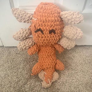 Può includere: Un giocattolo di peluche axolotl lavorato a uncinetto con filato arancione e beige. Il giocattolo ha un viso sorridente con occhi neri e una lunga coda arricciata.