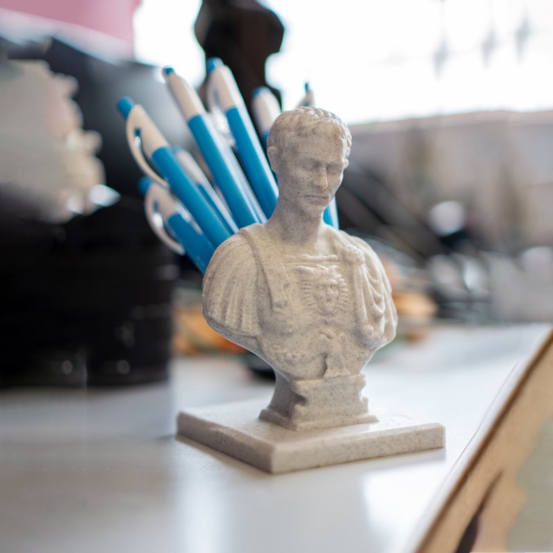 May include: A white bust of a man with a blue and white patterned shirt, used as a pen holder.  The bust is sitting on a white base and is surrounded by blue pens.