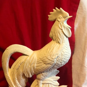 Puede incluir: Figura de gallo de cerámica blanca con un diseño esculpido detallado. El gallo está de pie sobre una base con hojas y un objeto redondo.