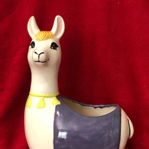May include: White ceramic llama planter with a purple and yellow saddle. The llama has a yellow mane and black eyes.