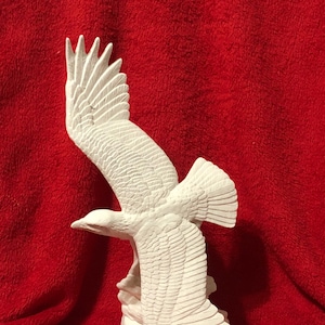 May include: White ceramic sculpture of an eagle in flight, perched on a rock. The eagle's wings are spread wide, and its talons are extended. The sculpture is detailed with feathers and other anatomical features.