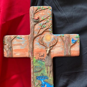 Puede incluir: Una cruz de madera con una escena pintada de un bosque con un ciervo, pájaros y un conejo. El texto "God Loves All Creatures Great and Small" está escrito en la cruz.