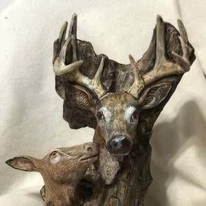 Puede incluir: Una escultura detallada de un ciervo y una cierva, tallada en un tronco de árbol. El ciervo tiene grandes astas y la cierva se acurruca contra el ciervo. La escultura está pintada en tonos marrones, con acentos blancos en las caras de los ciervos.