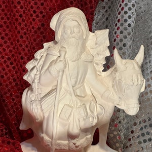 May include: White ceramic figurine of Santa Claus riding a donkey. Santa is holding a sack of gifts and a staff. The donkey is decorated with a Christmas tree.