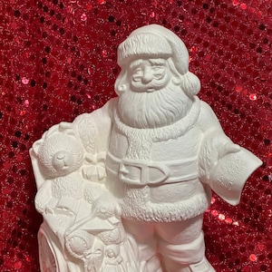 Könnte beinhalten: Weiße Keramikfigur vom Weihnachtsmann, der einen Teddybär hält und einen Wagen voller Spielzeug zieht. Der Wagen hat ein großes Rad und ein kleineres Rad hinten. Die Figur steht auf einem roten, glitzernden Hintergrund.