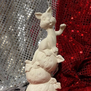 Könnte beinhalten: Weiße Keramikfigur eines Katers, der auf zwei Kürbissen sitzt. Die Kürbisse sind mit Herzen verziert. Die Figur steht auf einer roten Oberfläche.