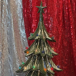 Può includere: Un albero di Natale in ceramica con finitura verde lucido. L'albero è adornato con luci colorate a forma di stella e una stella in cima. L'albero ha un design retrò classico, perfetto per le decorazioni natalizie.