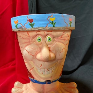 Può includere: Un vaso da fiori originale a forma di viso sorridente con pelle screpolata e occhi blu dipinti. Il vaso ha un bordo blu con fiori dipinti e poggia su due piedi.