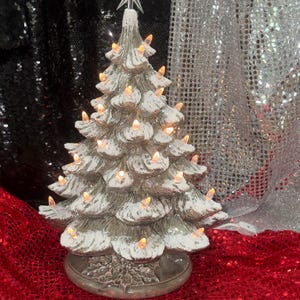 Könnte beinhalten: Ein weihnachtlicher Tischbaum aus Keramik mit glänzend grauer Oberfläche und weißer, schneeartiger Textur auf den Zweigen. Der Baum wird von kleinen, warm leuchtenden Lichtern erhellt und ist mit einem klaren Kunststoffstern gekrönt.