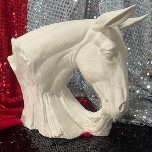 May include: A white horse head sculpture with detailed facial features and a flowing mane. The sculpture is made of a smooth, matte material and is set against a backdrop of red, silver, and black sequined fabric.