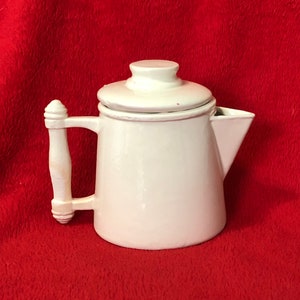 May include: A white ceramic coffee pot with a lid and a handle. The pot has a spout for pouring and sits on a red surface. The handle is a decorative design.