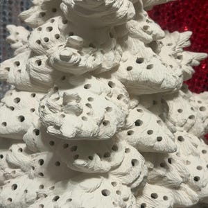 Pu&ograve; includere: Primo piano di un albero di Natale in ceramica bianca con una superficie strutturata e porosa. L'albero ha un design dettagliato e stratificato con numerosi piccoli fori, creando un aspetto decorativo unico. L'albero &egrave; non verniciato e pronto per la personalizzazione.