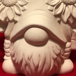 May include: White ceramic gnome figurine with a beard and a hat made of sunflowers. The gnome has a round nose and is standing on a red surface.
