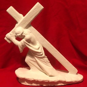 May include: A white ceramic sculpture depicting a figure carrying a large cross. The figure is draped in flowing robes and appears to be kneeling. The sculpture is set on a cloud-like base against a red background.