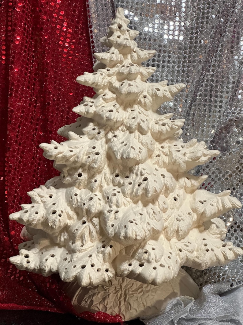 Pu&ograve; includere: Un albero di Natale in ceramica color crema con una superficie strutturata e numerosi piccoli fori. L'albero ha un design dettagliato e stratificato e poggia su una base decorativa. Lo sfondo presenta un tessuto con paillettes rosse e argentate.