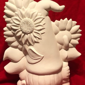 May include: White ceramic gnome figurine with a sunflower design. The gnome is wearing a hat and holding a sunflower in its hand.