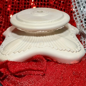 Può includere: Un porta torta in ceramica bianca con base a forma di stella e parte superiore rotonda. Il porta torta è decorato con un design in rilievo di linee e curve.