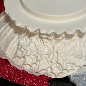 Pu&ograve; includere: Un pezzo decorativo bianco, con bordo smerlato, con un foro centrale, forse per un puntale per albero di Natale. Il pezzo ha un design in rilievo strutturato di foglie di agrifoglio. La base &egrave; stratificata con tessuto glitterato rosso e paillettes nere.