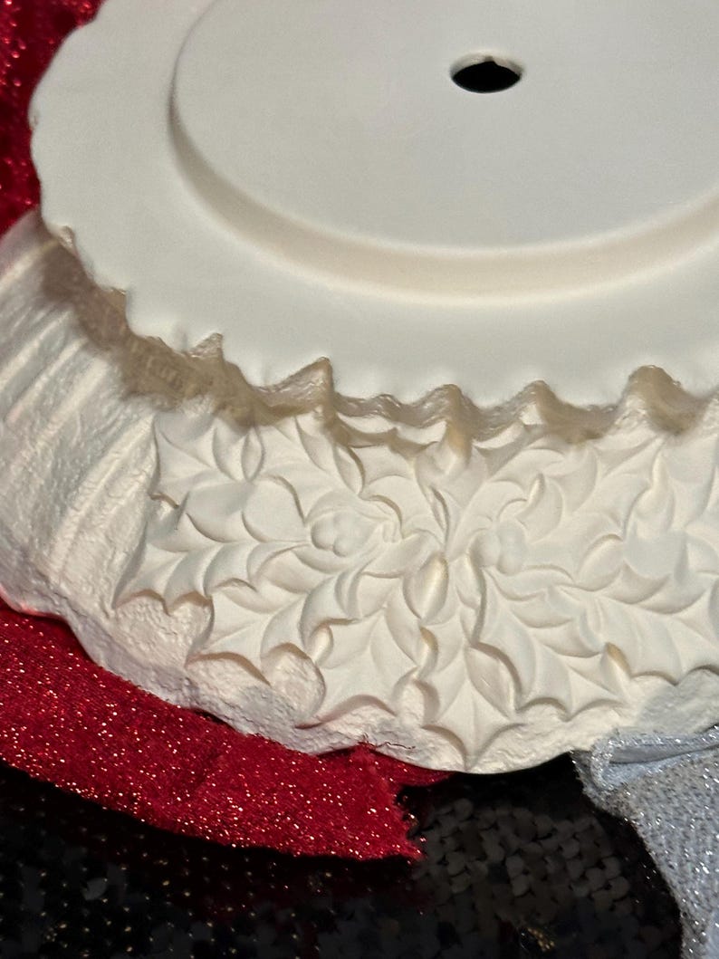 Pu&ograve; includere: Un pezzo decorativo bianco, con bordo smerlato, con un foro centrale, forse per un puntale per albero di Natale. Il pezzo ha un design in rilievo strutturato di foglie di agrifoglio. La base &egrave; stratificata con tessuto glitterato rosso e paillettes nere.