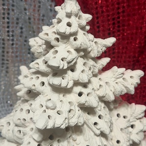 Pu&ograve; includere: Un albero di Natale in ceramica bianca con una superficie strutturata e numerosi piccoli fori. L'albero ha un design dettagliato e stratificato ed &egrave; probabilmente destinato a scopi decorativi. Lo sfondo presenta un mix di trame argentate e rosse scintillanti.