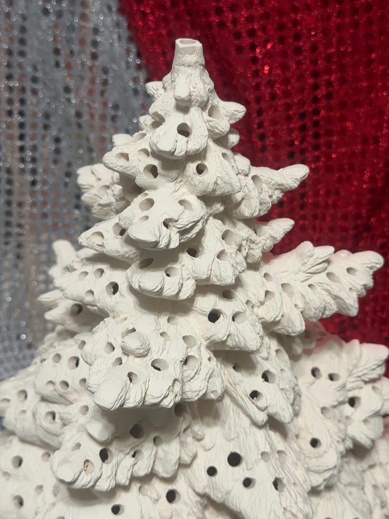 Pu&ograve; includere: Un albero di Natale in ceramica bianca con una superficie strutturata e numerosi piccoli fori. L'albero ha un design dettagliato e stratificato ed &egrave; probabilmente destinato a scopi decorativi. Lo sfondo presenta un mix di trame argentate e rosse scintillanti.