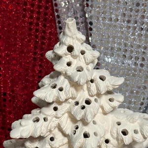 Pu&ograve; includere: Un albero di Natale in ceramica bianca con una superficie strutturata e numerosi piccoli fori. L'albero ha la forma tradizionale di un abete ed &egrave; progettato per contenere una luce all'interno. Lo sfondo &egrave; rosso e argento.