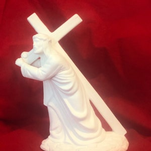 May include: White ceramic figurine of a person carrying a cross on their shoulders. The figurine is in a kneeling position with the cross resting on their back. The figurine is on a white base.