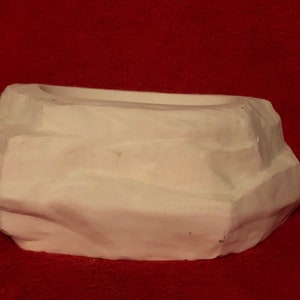 Könnte beinhalten: Weißer Keramik-Pflanztopf in Form eines Steins mit einer glatten, abgerundeten Oberfläche. Der Topf hat eine flache, ovale Öffnung oben.