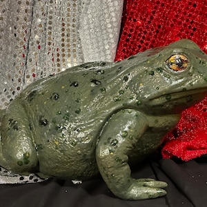 May include: A green toad figurine with black spots and a textured surface. The toad is sitting on a black surface with a silver and red sparkly background.