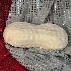 Puede incluir: Un plato de cerámica blanca con forma de cacahuete. El plato está sobre una tela brillante plateada y roja.