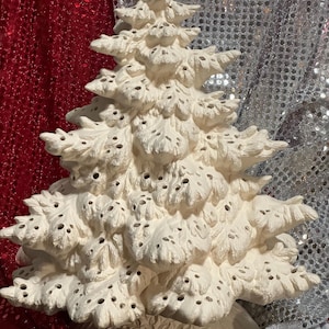 Pu&ograve; includere: Un albero di Natale in ceramica color crema con una superficie strutturata e porosa. L'albero ha un design dettagliato e stratificato e poggia su una base decorativa. Lo sfondo presenta un tessuto di paillettes rosse e argentate.