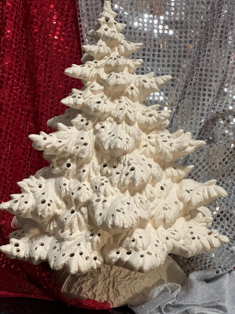 Pu&ograve; includere: Un albero di Natale in ceramica color crema con una superficie strutturata e porosa. L'albero ha un design dettagliato e stratificato e poggia su una base decorativa. Lo sfondo presenta un tessuto di paillettes rosse e argentate.