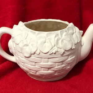 May include: A white ceramic teapot with a basket weave design on the base and floral accents around the rim. The teapot has a handle and a spout, perfect for a decorative piece or for painting.