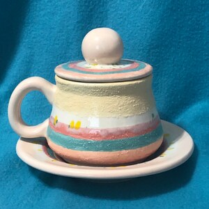 May include: A ceramic teacup and saucer set with a yellow, pink, and blue striped design and small yellow flower accents. The teacup has a lid with a round knob.