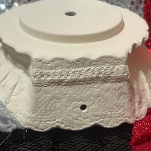 Pu&ograve; includere: Un piedistallo in ceramica bianca con bordo smerlato e foro centrale. Il piedistallo presenta un design in pizzo strutturato attorno alla base e una parte superiore circolare, adatto per esporre oggetti decorativi.