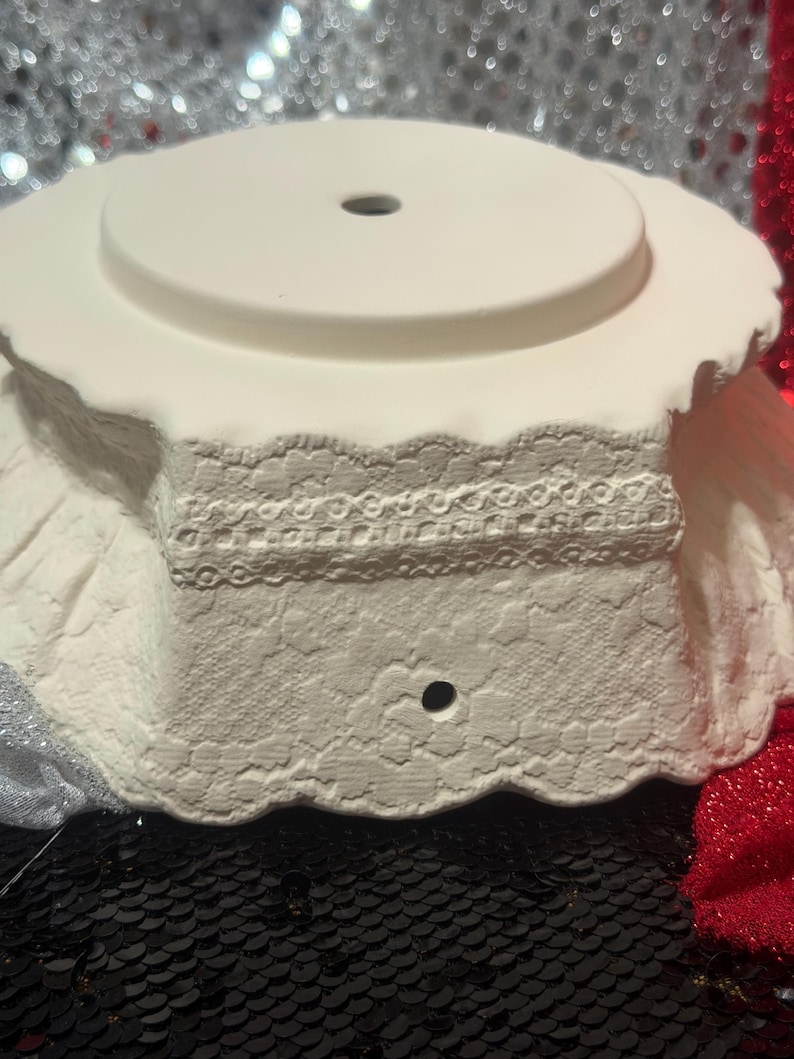 Pu&ograve; includere: Un piedistallo in ceramica bianca con bordo smerlato e foro centrale. Il piedistallo presenta un design in pizzo strutturato attorno alla base e una parte superiore circolare, adatto per esporre oggetti decorativi.