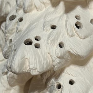 Pu&ograve; includere: Primo piano di una scultura in ceramica bianca strutturata con pi&ugrave; fori. La scultura ha una forma organica a strati con una superficie ruvida e irregolare. I fori sono scuri e uniformemente distanziati.