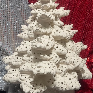 Pu&ograve; includere: Un albero di Natale in ceramica bianca con un design strutturato e stratificato. L'albero presenta numerosi piccoli fori nei suoi rami. Lo sfondo presenta tessuti di paillettes argentate e rosse.