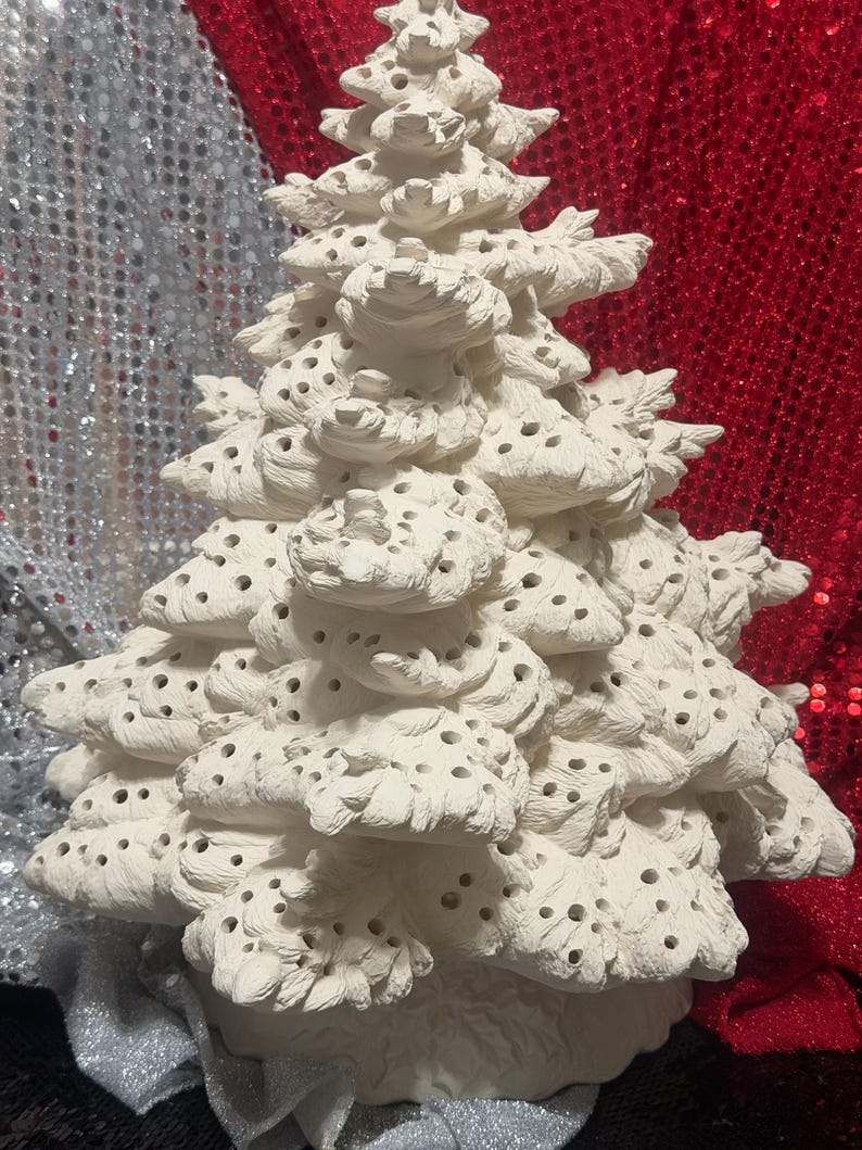 Pu&ograve; includere: Un albero di Natale in ceramica bianca con un design strutturato e stratificato. L'albero presenta numerosi piccoli fori nei suoi rami. Lo sfondo presenta tessuti di paillettes argentate e rosse.