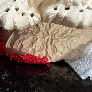 Pu&ograve; includere: Primo piano di un pezzo di ceramica strutturata color crema con pi&ugrave; fori, forse un oggetto decorativo. Un nastro rosso glitterato e un tessuto argentato sono visibili. Lo sfondo &egrave; una superficie nera con paillettes.