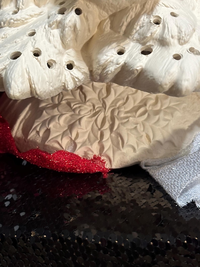 Pu&ograve; includere: Primo piano di un pezzo di ceramica strutturata color crema con pi&ugrave; fori, forse un oggetto decorativo. Un nastro rosso glitterato e un tessuto argentato sono visibili. Lo sfondo &egrave; una superficie nera con paillettes.