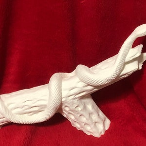 May include: White ceramic sculpture of a snake coiled around a tree branch. The snake is in a raised position with its head tilted upwards. The sculpture is unpainted and ready to be decorated.