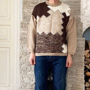 Puede incluir: Un suéter de color marrón y beige con cuello redondo. El suéter tiene un diseño geométrico con colores marrón, beige y blanco.