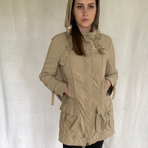 May include: A beige hooded jacket with multiple pockets and a zip fastening. The jacket has a tailored fit and is worn with black trousers and black boots. The jacket features decorative straps and a high collar.