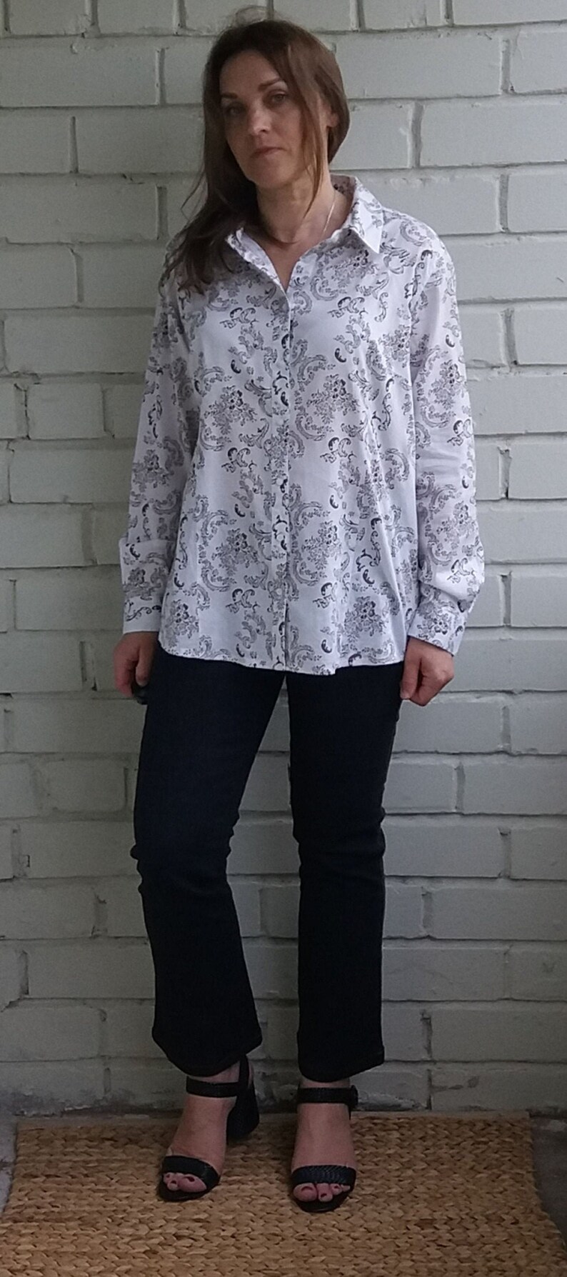 May include: A white long-sleeved shirt with a black floral pattern. The shirt has a button-up front and a collared neckline. The person is wearing black pants and black heeled sandals.